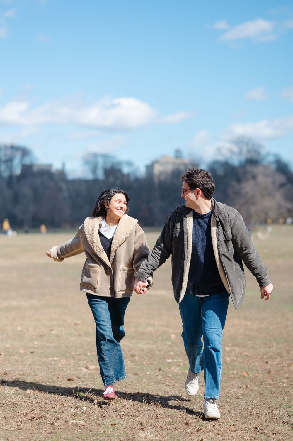 Two people in casual attire playfully run and chase each other across an open field on a sunny day.