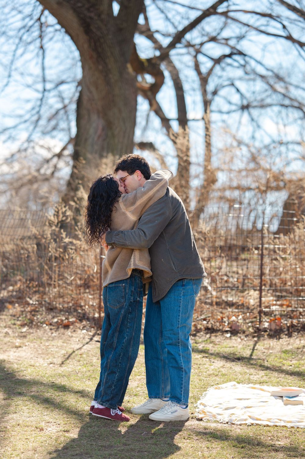 A couple shares a romantic kiss on a sunny winter day in a park with bare trees in the background in Prospect Park in Brooklyn.