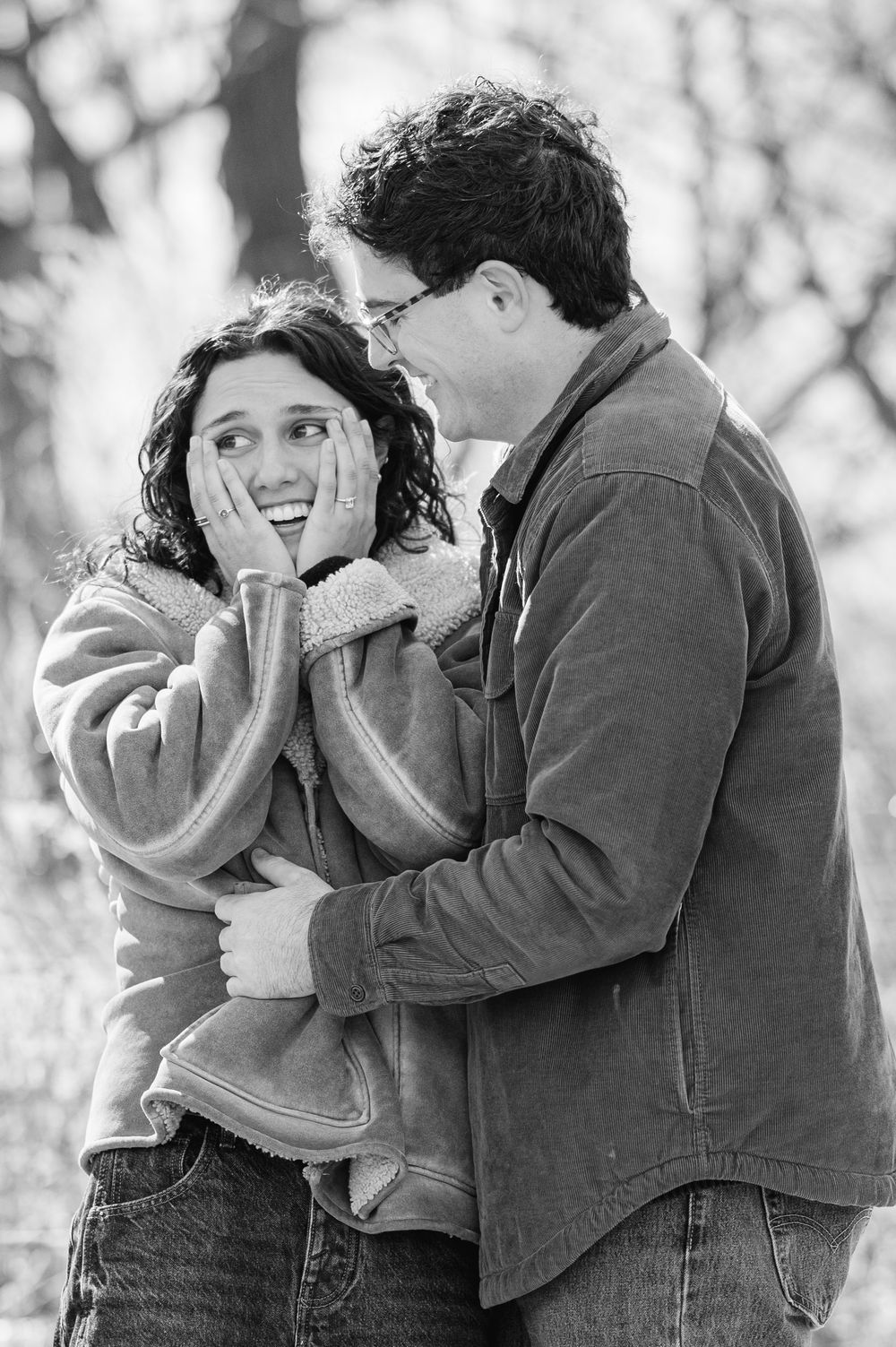 A black and white candid moment of a shocked new fianceé clutching her face in surprise right after she gets engaged with her fiance smiling