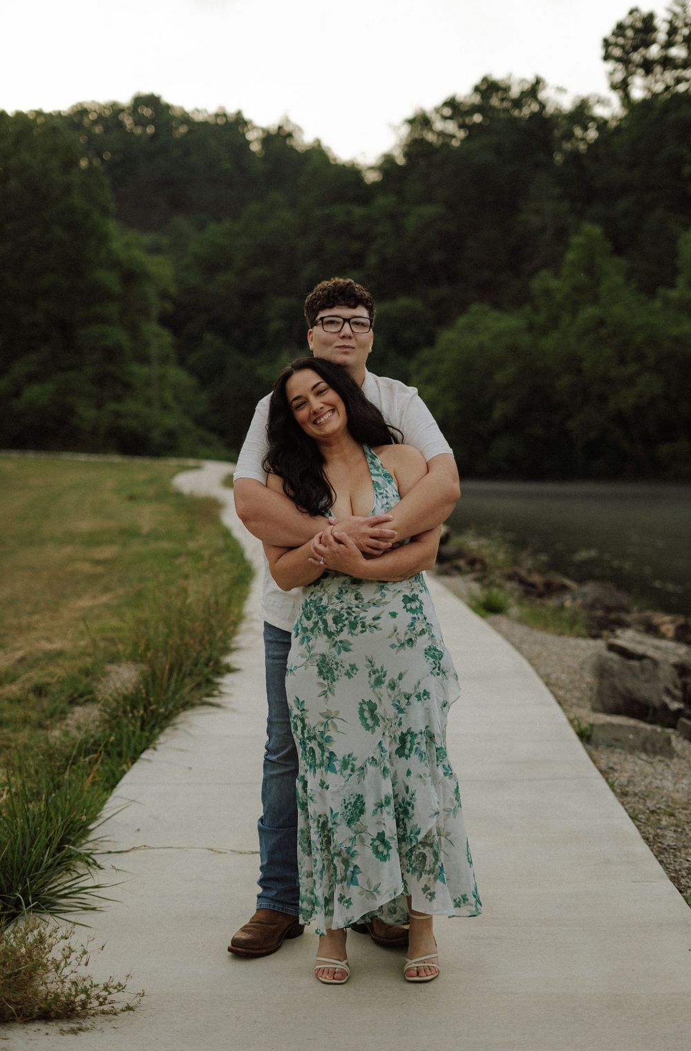 Couple embracing on a lakeside path, woman in floral dress, man in white shirt and jeans, lush green trees behind.