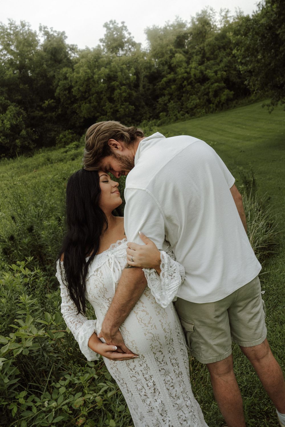 Couple sharing a tender moment in a lush green field, woman in white lace maternity dress, man in white shirt and khaki shorts.