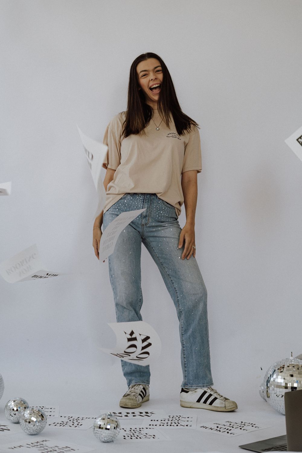 Young woman laughing in beige tee, jeans, and Adidas sneakers surrounded by disco balls and confetti.