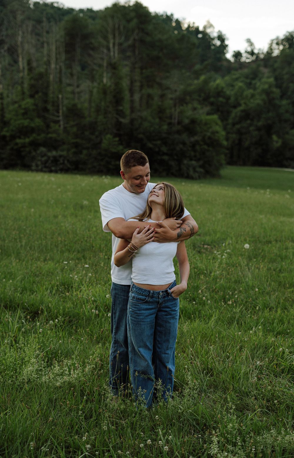 Couple embracing in a lush green meadow, wearing white tops and jeans, with dense forest in the background.