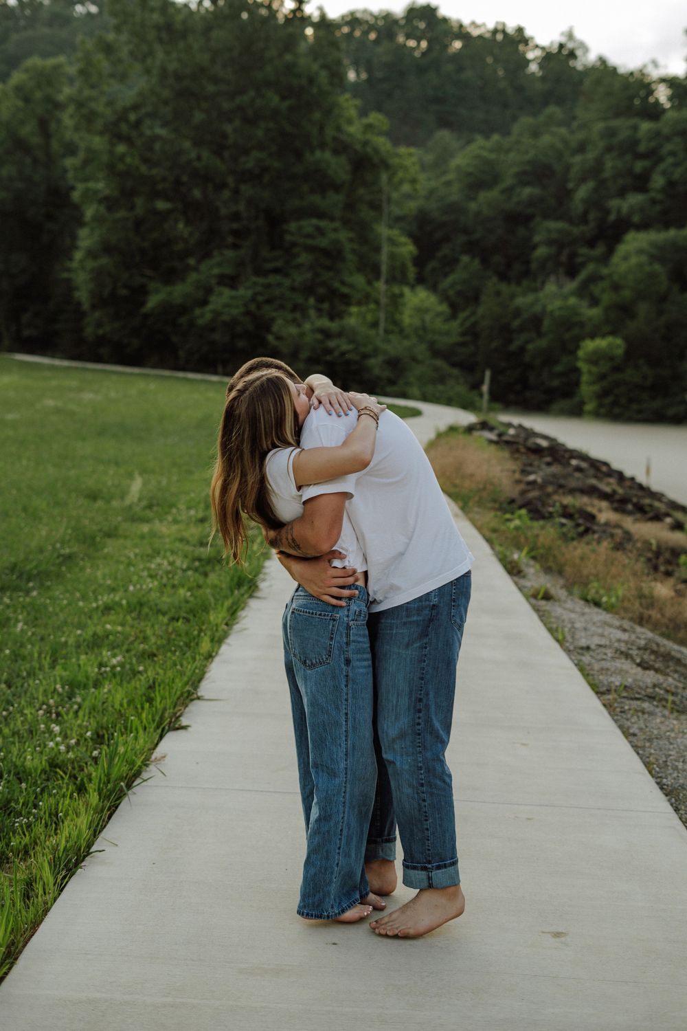 Two people sharing a warm embrace on a concrete path surrounded by lush green trees and grass.