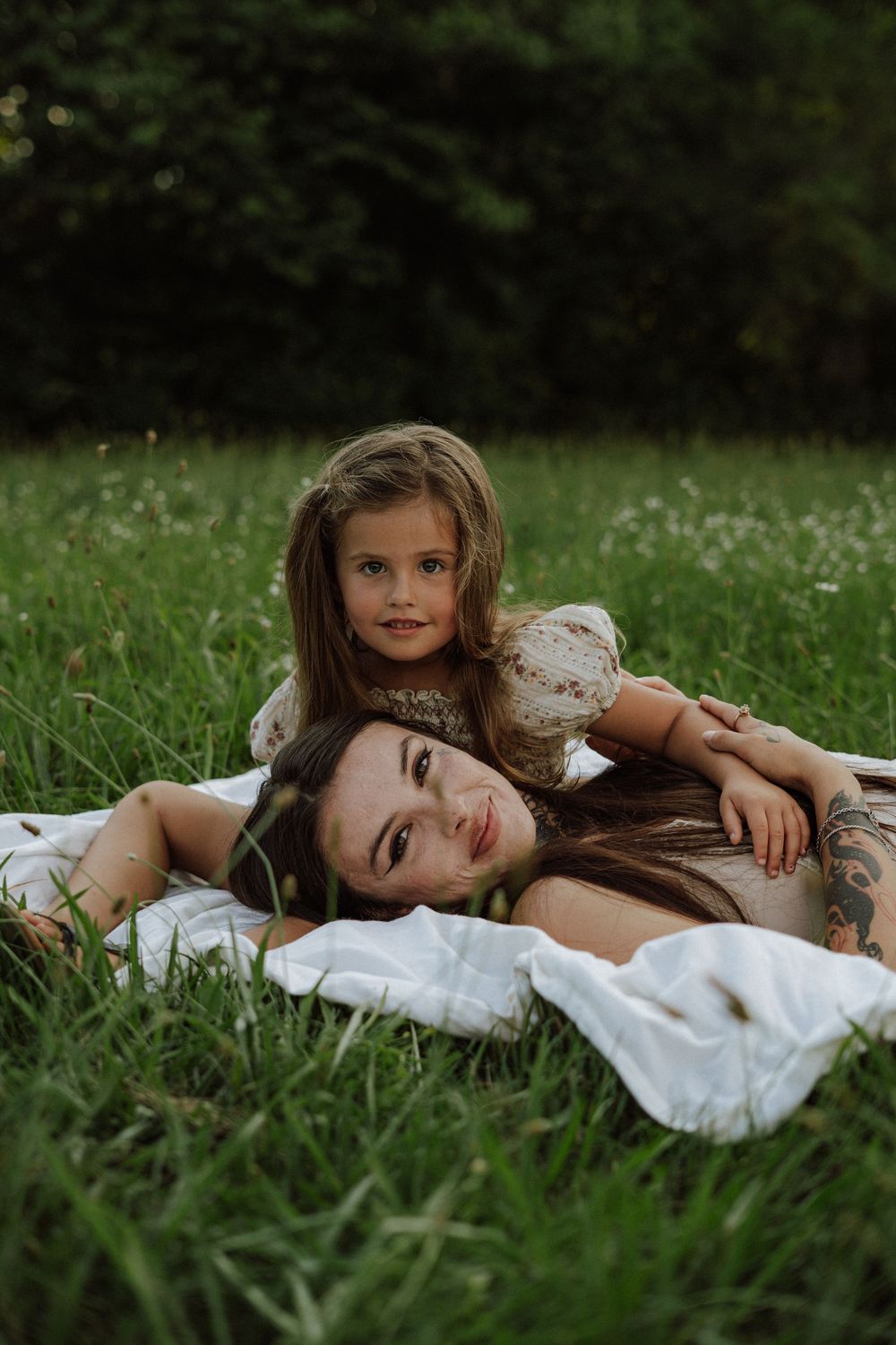 A young girl rests her chin on a woman lying on a white blanket in lush green grass outdoors.