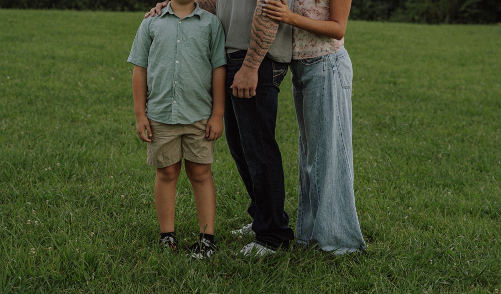 Family of three standing on green grass, showing torsos and legs, boy in green shirt and khaki shorts.