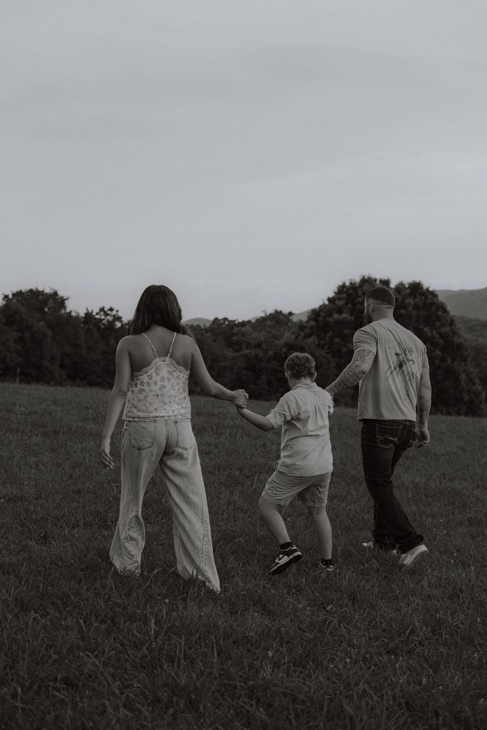 Black and white photo of a family holding hands walking through a green meadow with trees in the background.