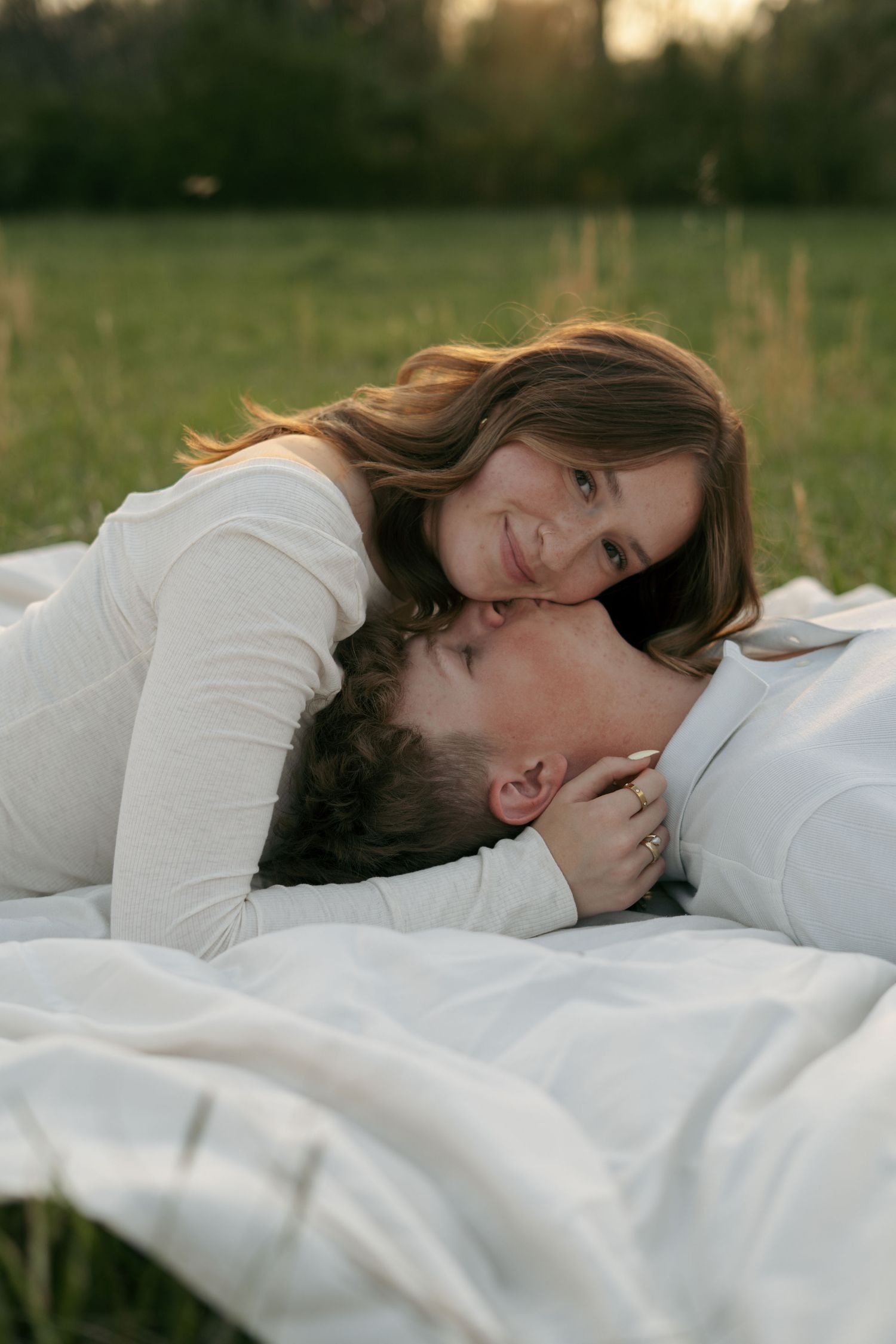 A smiling couple lying on a white blanket in a green field, sharing a tender romantic moment outdoors.