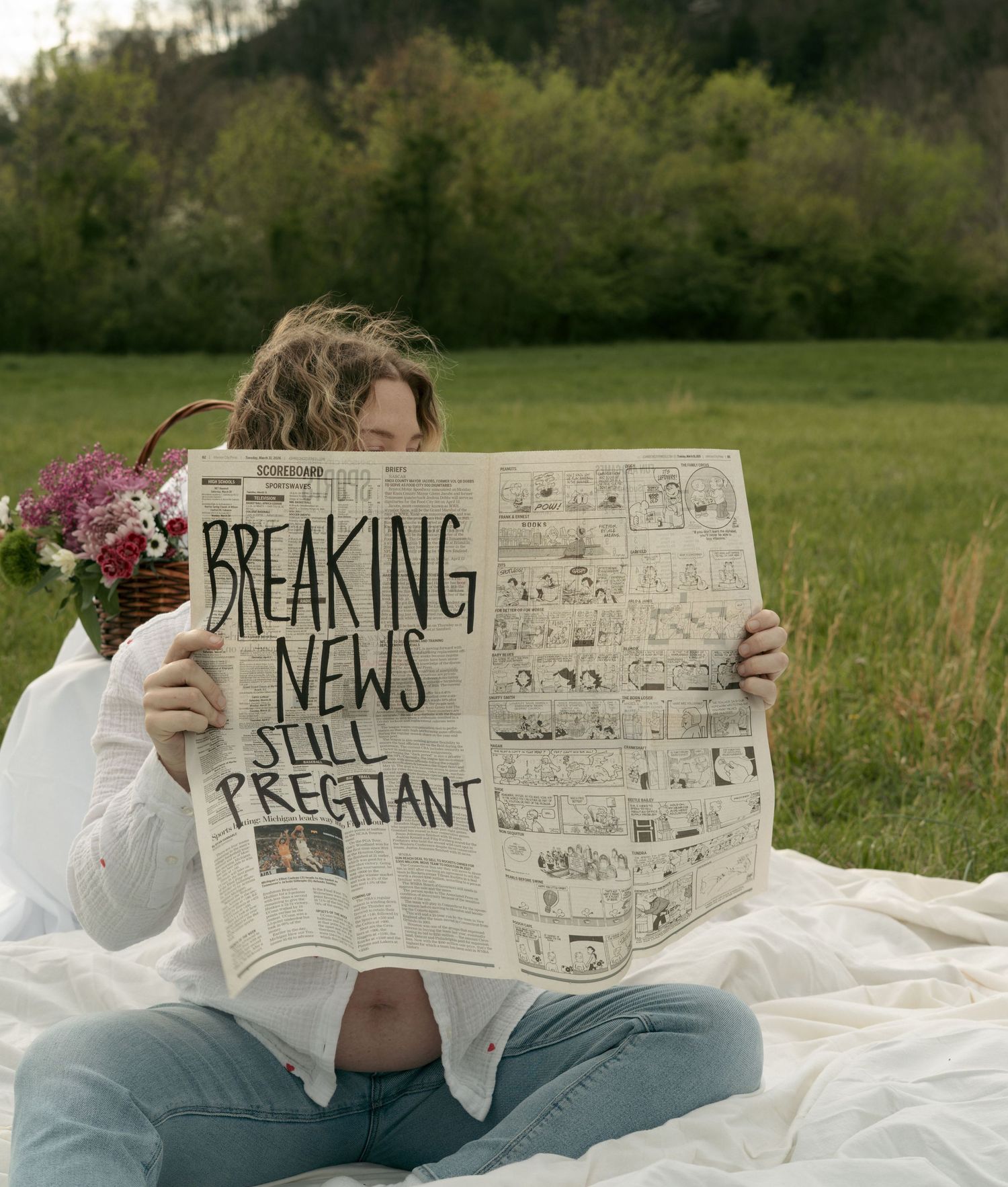 Pregnant woman sitting outdoors on white blanket holding newspaper with humorous Breaking News Still Pregnant headline.