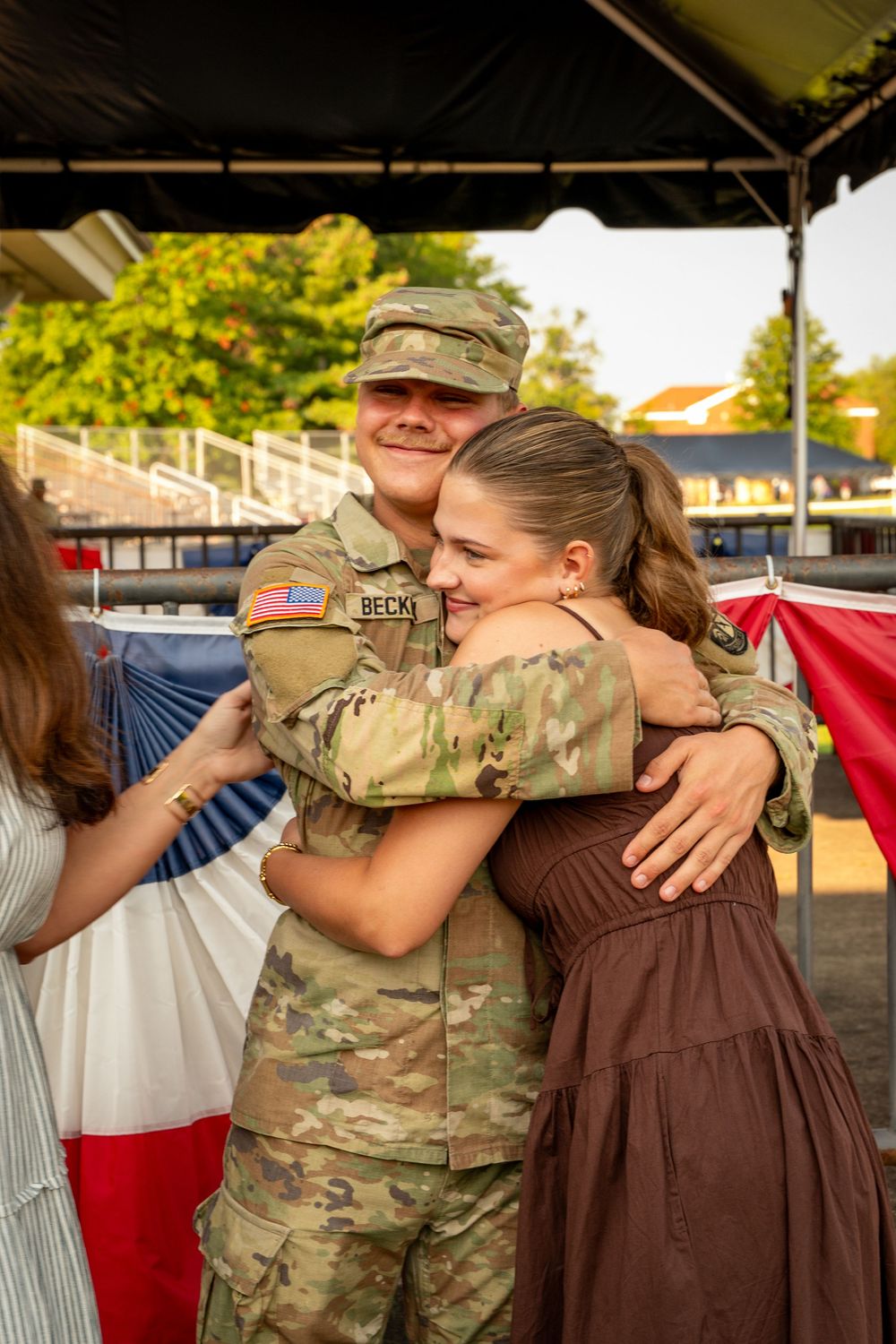Military service member in camouflage uniform embraces loved one during emotional outdoor gathering.
