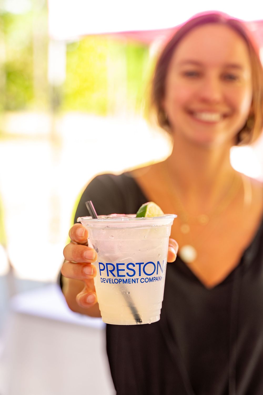 A hand holds up a clear plastic cup with a "Preston" logo containing a refreshing beverage with lime garnish.
