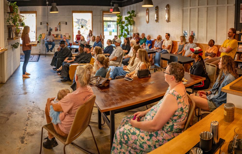 People gather in a modern community space for a presentation or meeting with wooden benches and bright lighting.