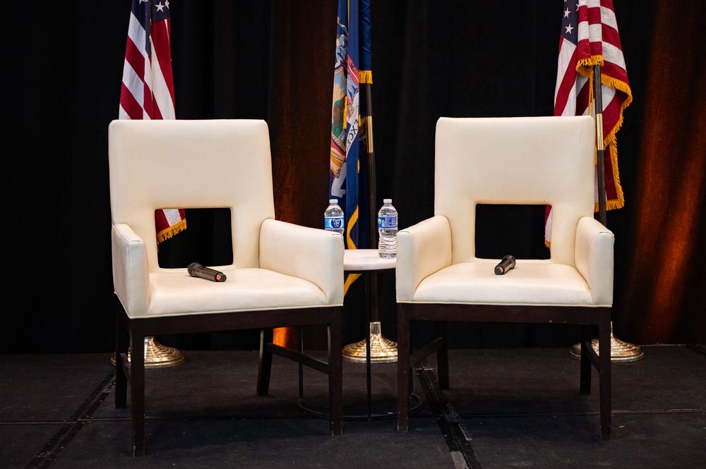 A stage setup with white armchairs and American flags displayed behind them during a formal event.
