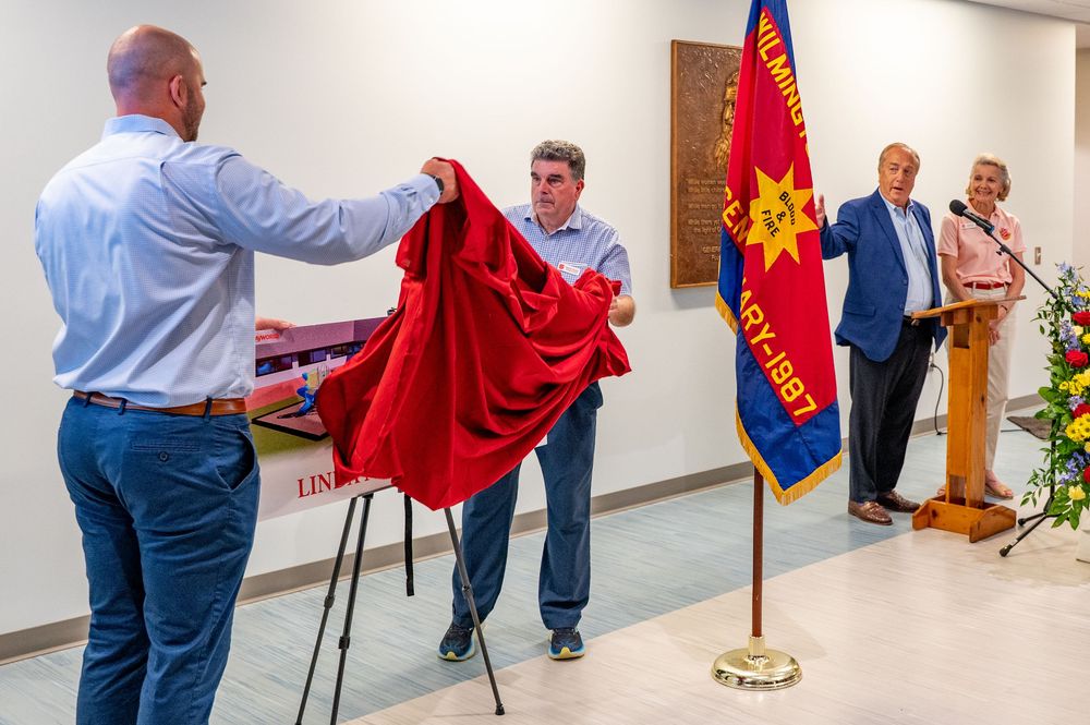 People prepare ceremonial items and flags for an event in a hallway with a military or organizational banner displayed.
