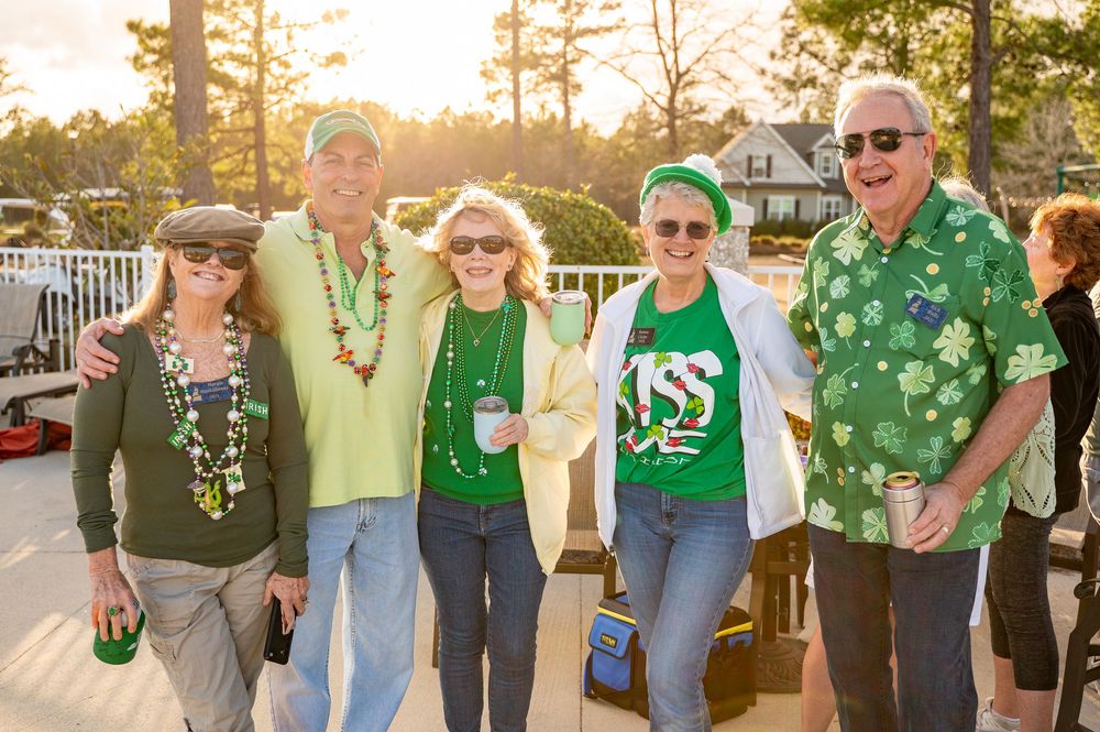 A group of people wearing festive green clothing and accessories celebrate St. Patrick's Day outdoors at sunset.