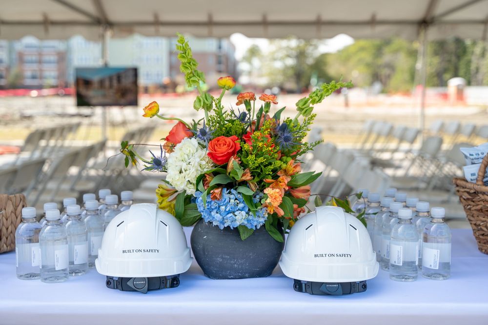 A colorful floral centerpiece with construction hard hats displayed on a table with water glasses at an outdoor event.