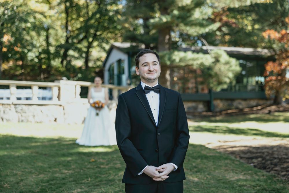 A person in a black tuxedo stands outdoors in a garden setting during a formal event.