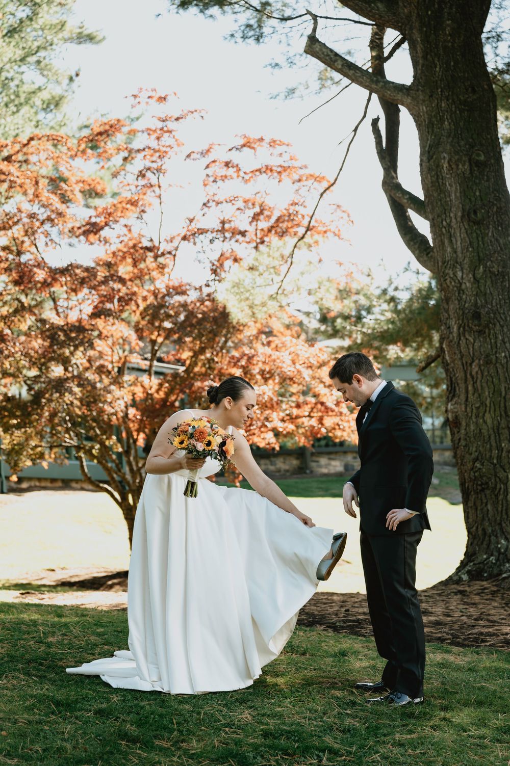 A couple in wedding attire stands beneath a blooming autumn tree with orange leaves during an outdoor ceremony.