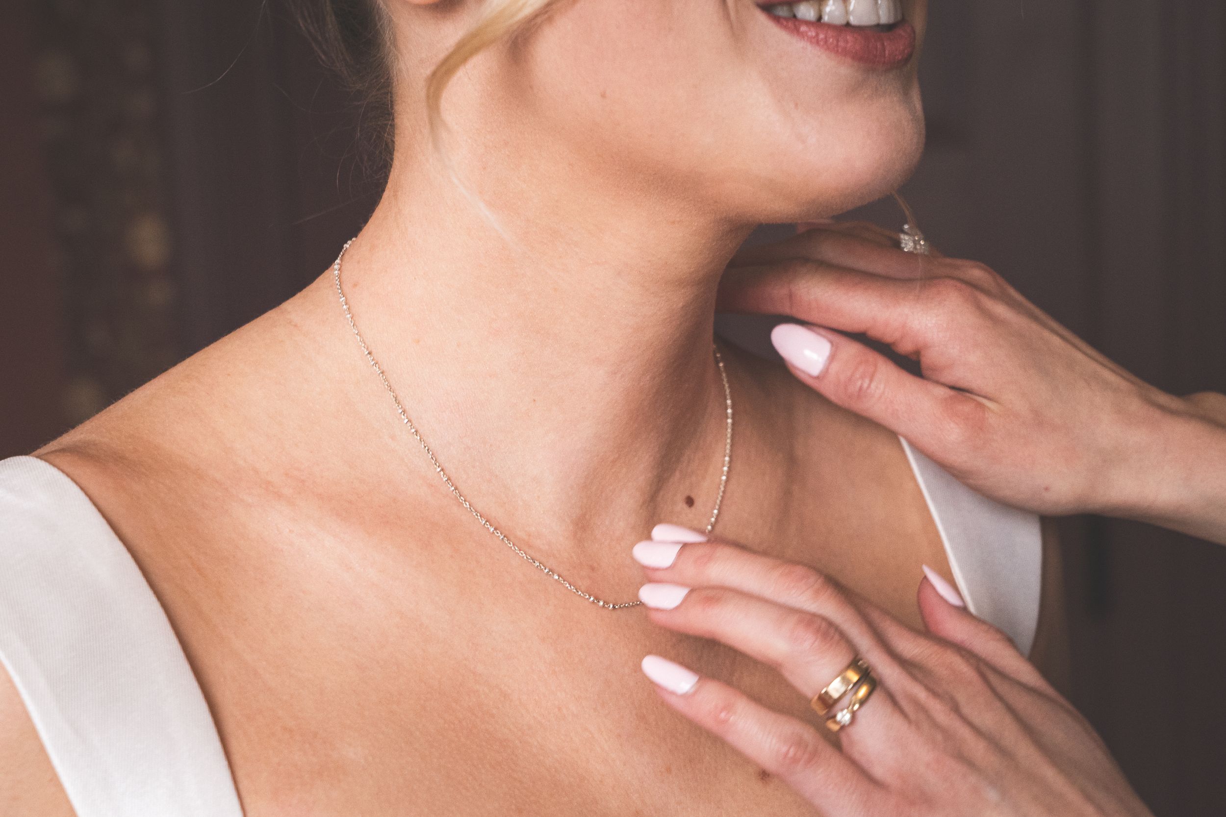 Close-up of a smiling bride wearing a delicate gold necklace, with manicured hands adjusting the jewelry.