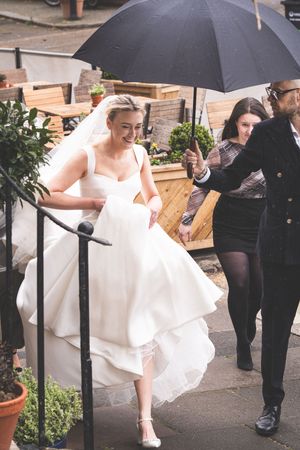 Bride in white gown ascending steps with helper holding black umbrella on a rainy wedding day.