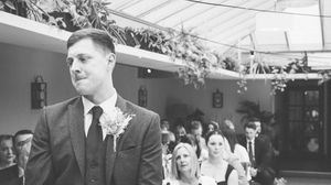 Groom in grey suit with boutonniere awaiting bride at wedding ceremony in elegant venue.