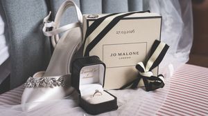 Wedding flat lay featuring Jo Malone gift box, diamond ring in box, and bridal heels on white fabric.
