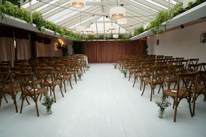 Indoor wedding ceremony space with wooden chairs arranged in rows under a greenhouse-style ceiling with hanging greenery.