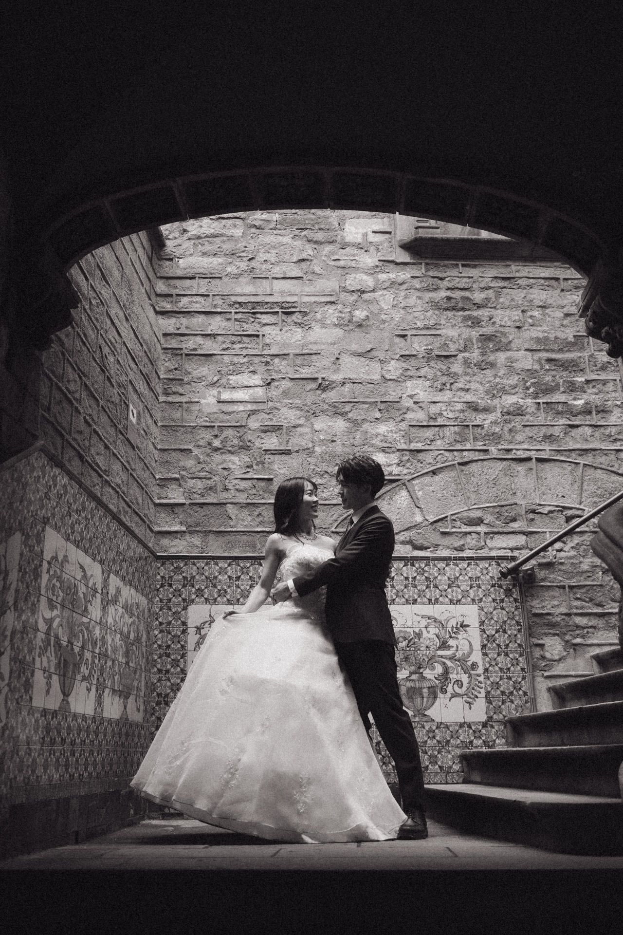 A romantic black and white photo of a couple in wedding attire dancing under a stone archway with stairs in the background.