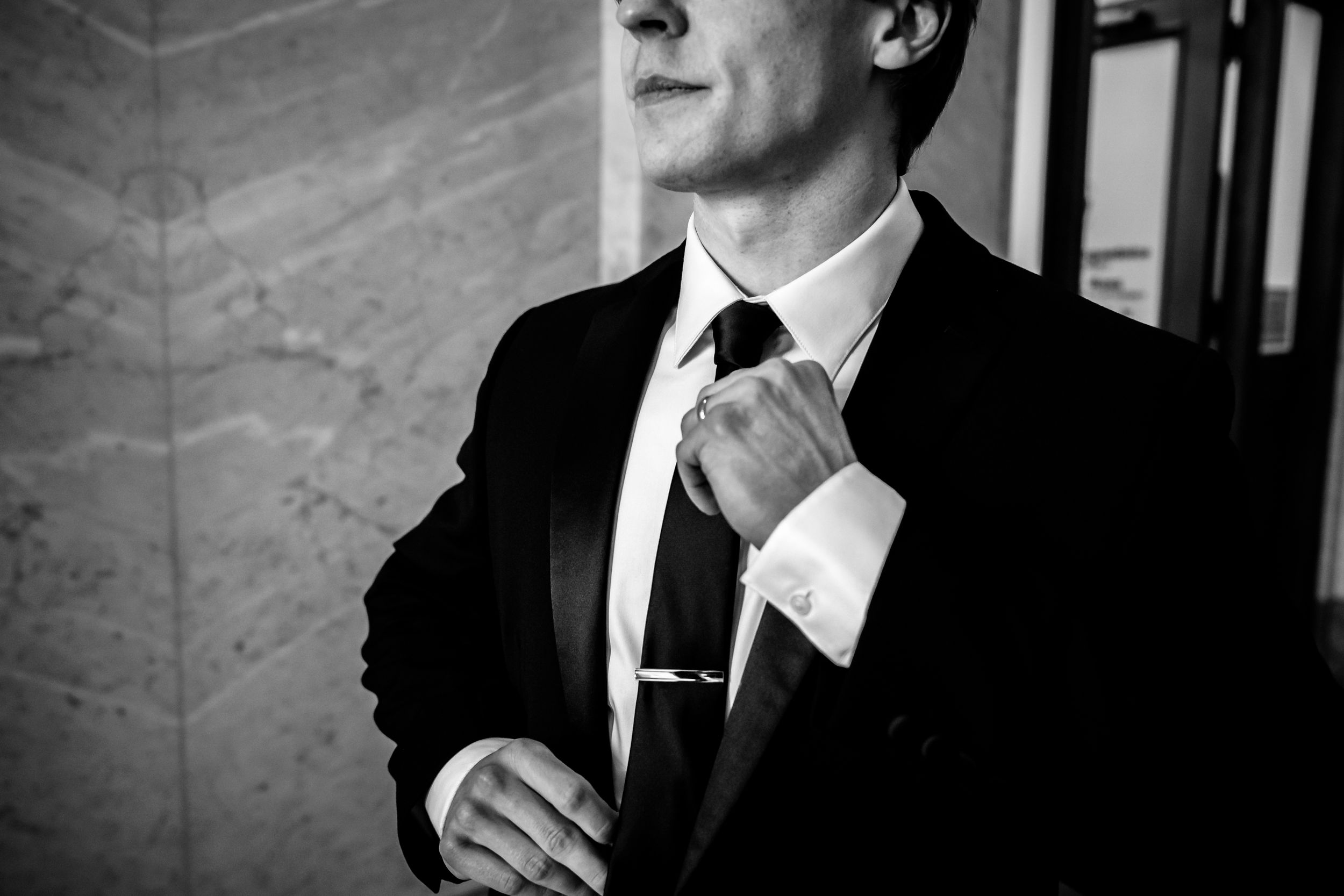 A man in a black suit adjusts his tie while looking away from the camera.