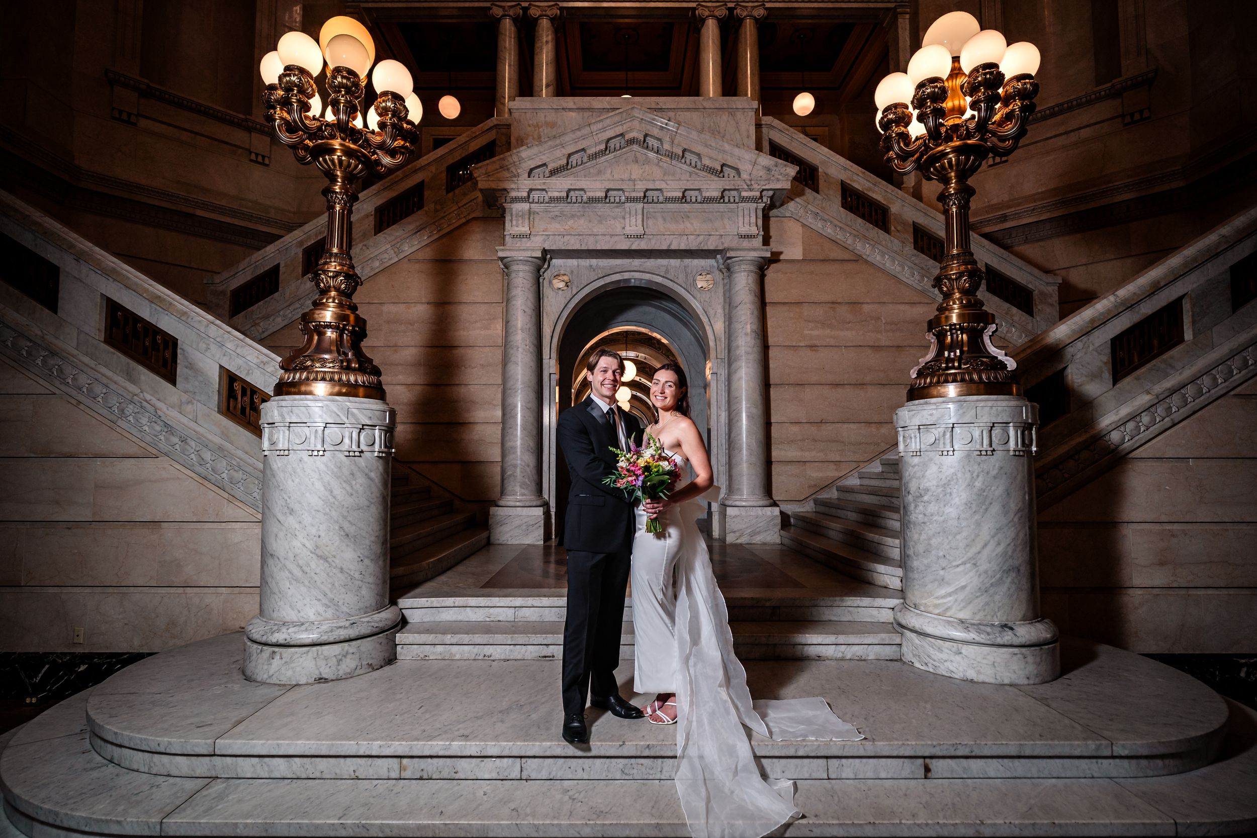 Elegant wedding couple poses on grand marble staircase with ornate chandeliers in historic building.