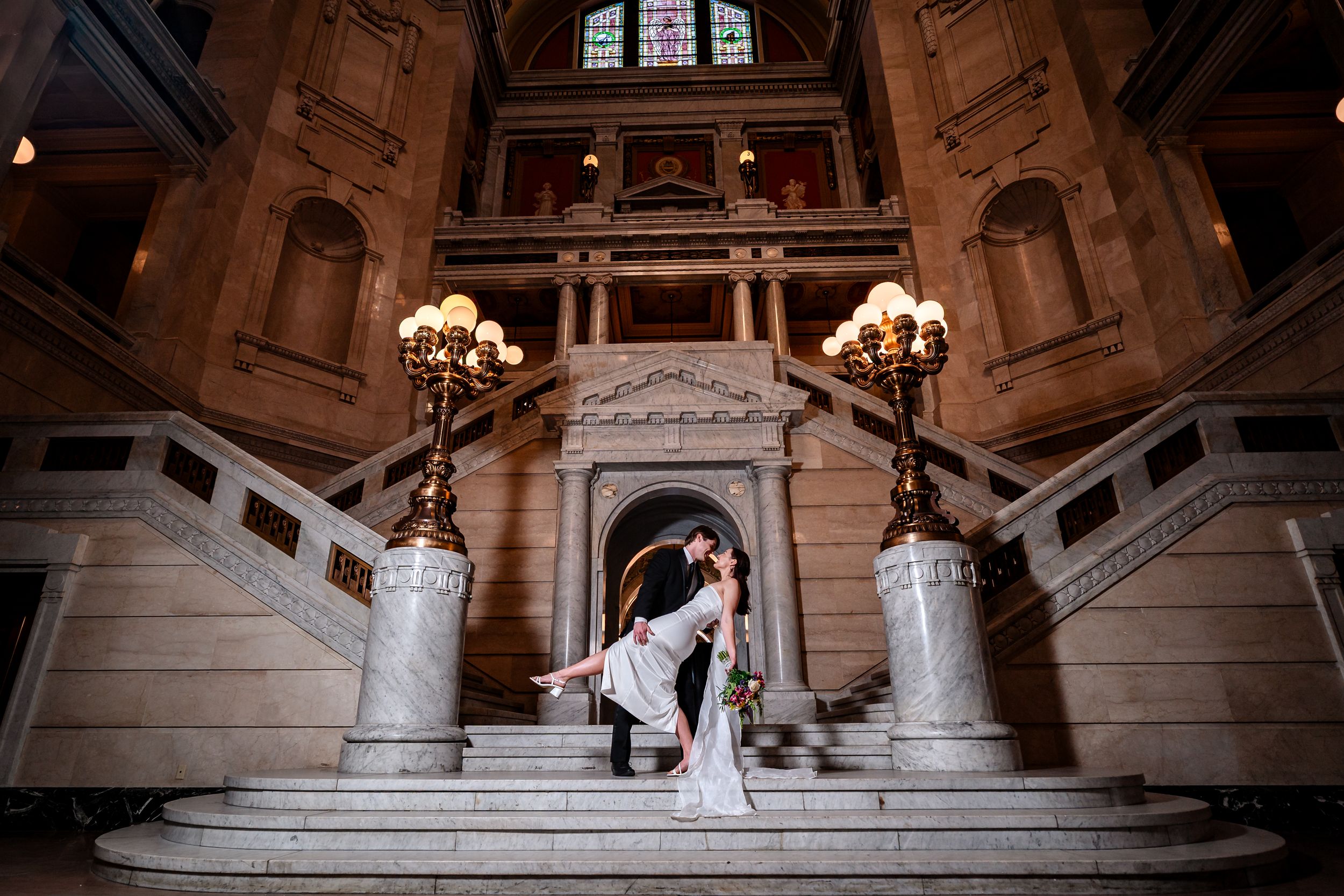 A couple shares a romantic moment on a grand marble staircase in an ornate historic building.