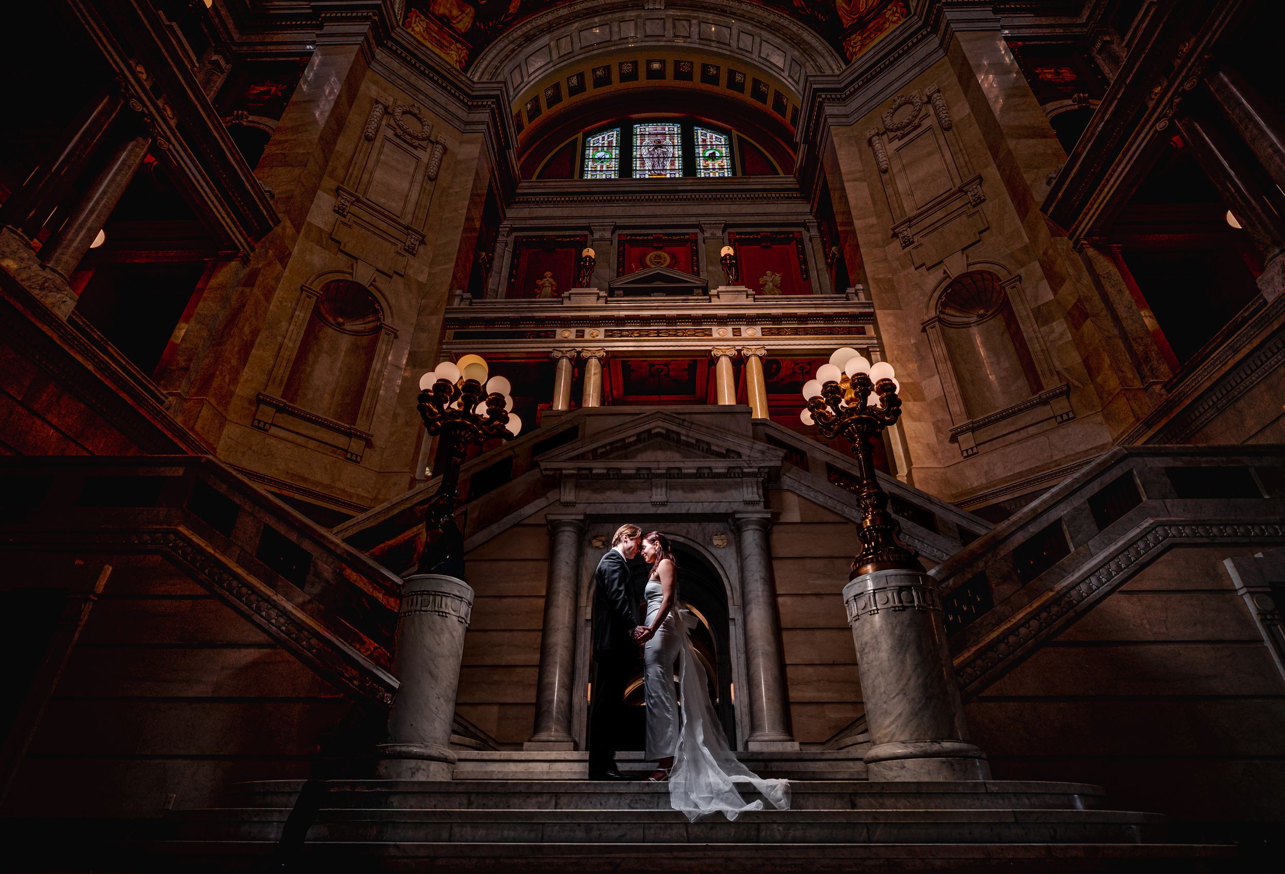 Elegant couple stands in dramatic archway of historic building with ornate architecture and warm lighting.