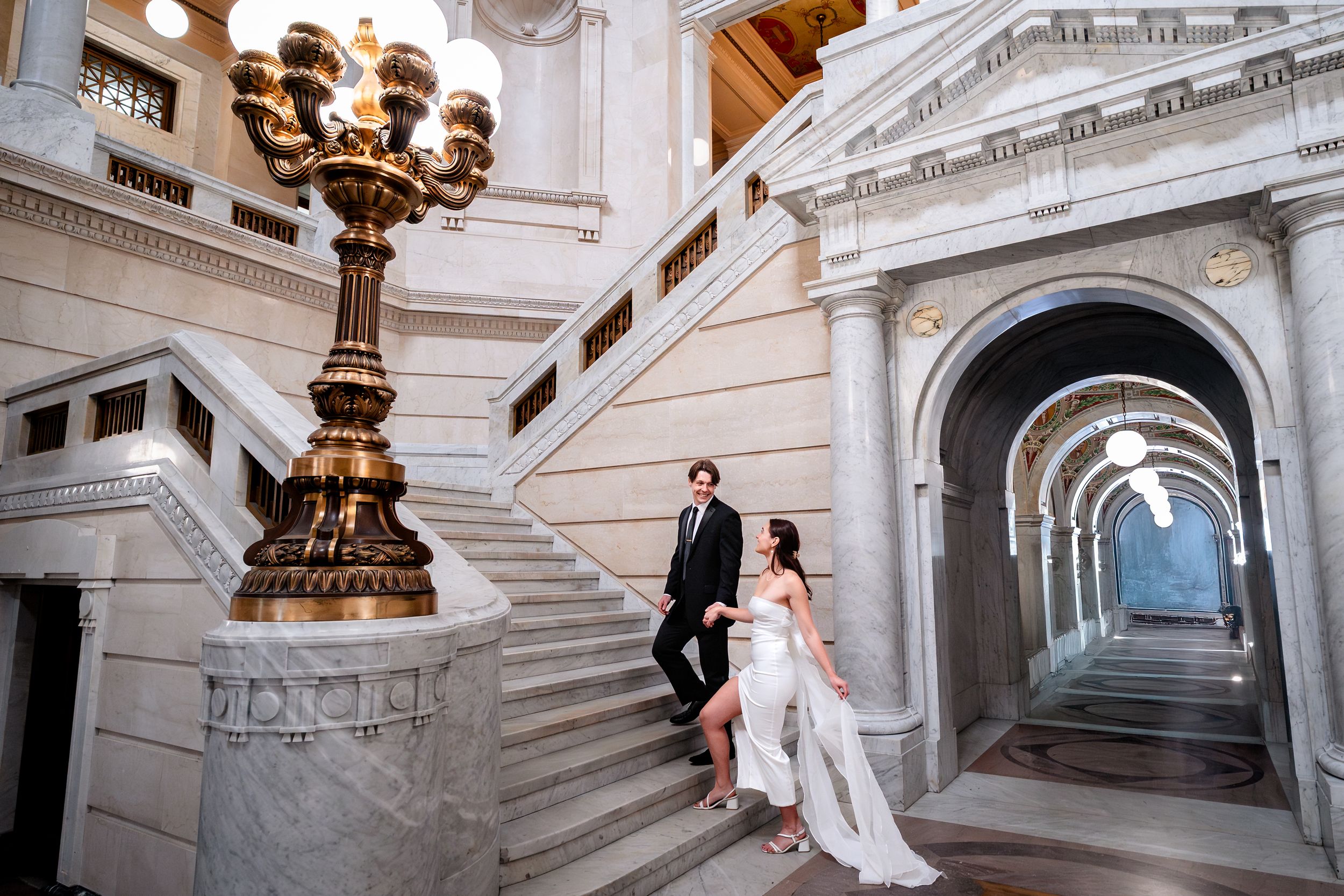 Elegant wedding couple poses on grand marble staircase in ornate historic building with classical architecture.