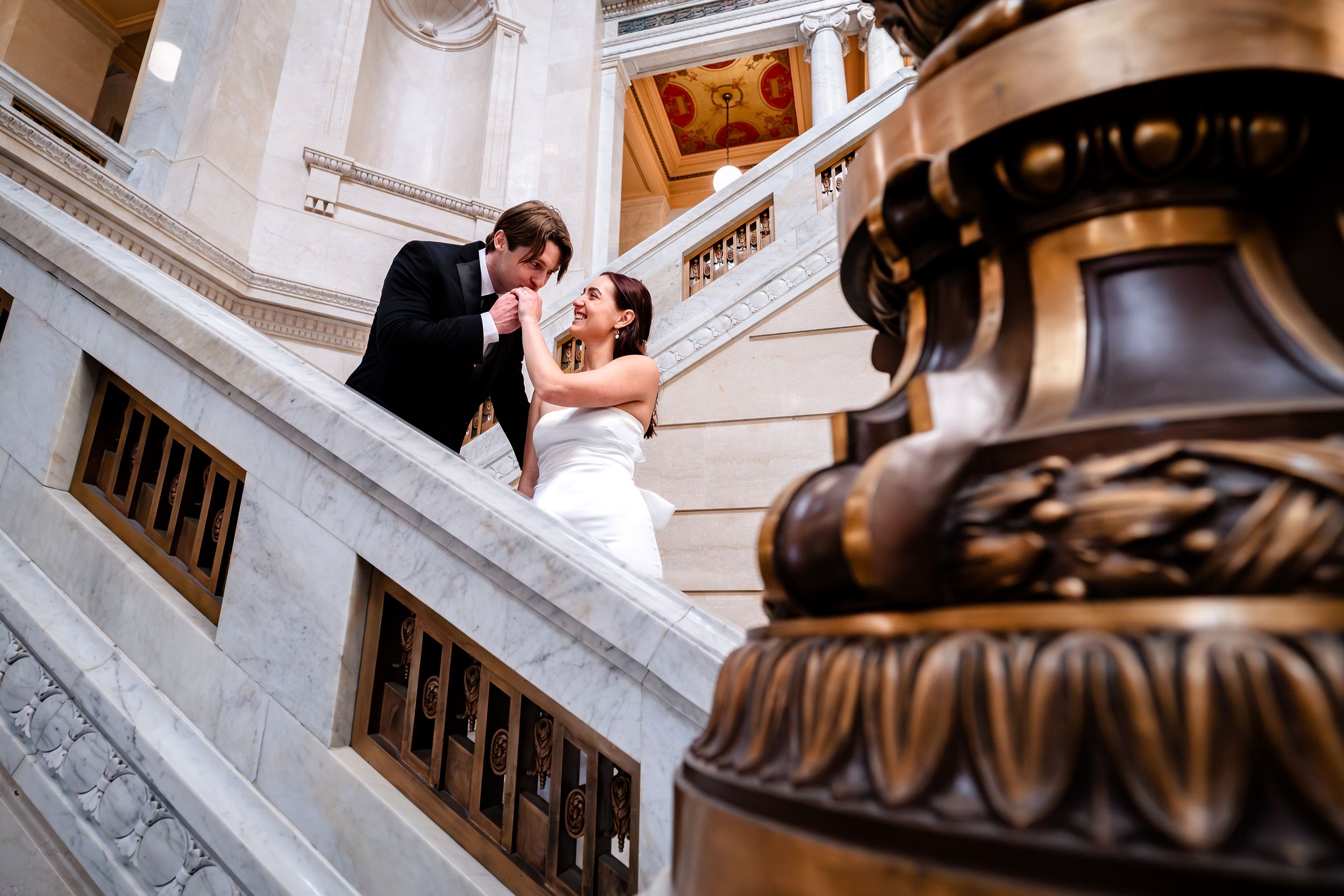 A couple shares a romantic moment on an elegant marble staircase with ornate architectural details.