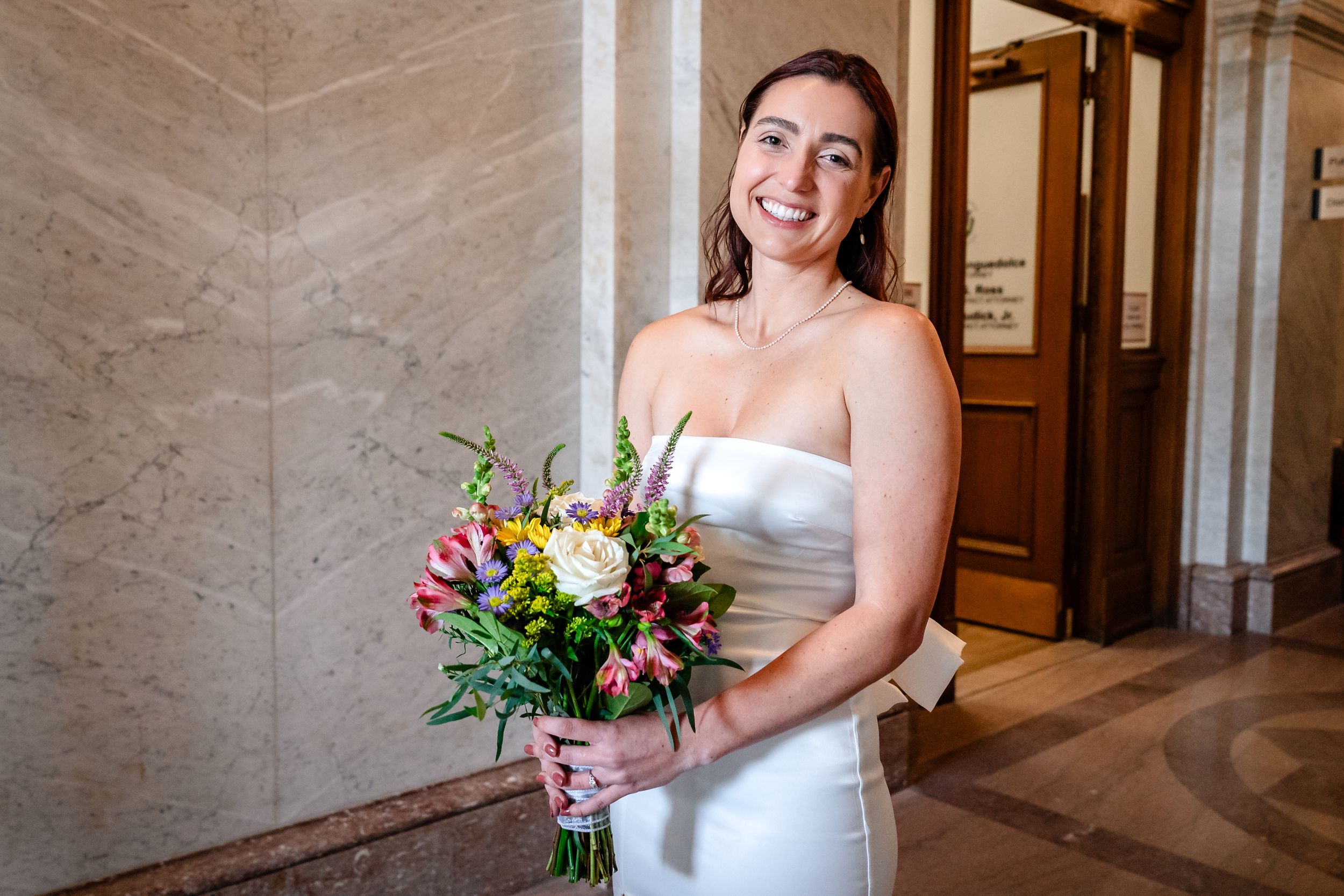 A bride in a white strapless gown holds a vibrant pink and yellow floral bouquet while standing in an elegant hallway.