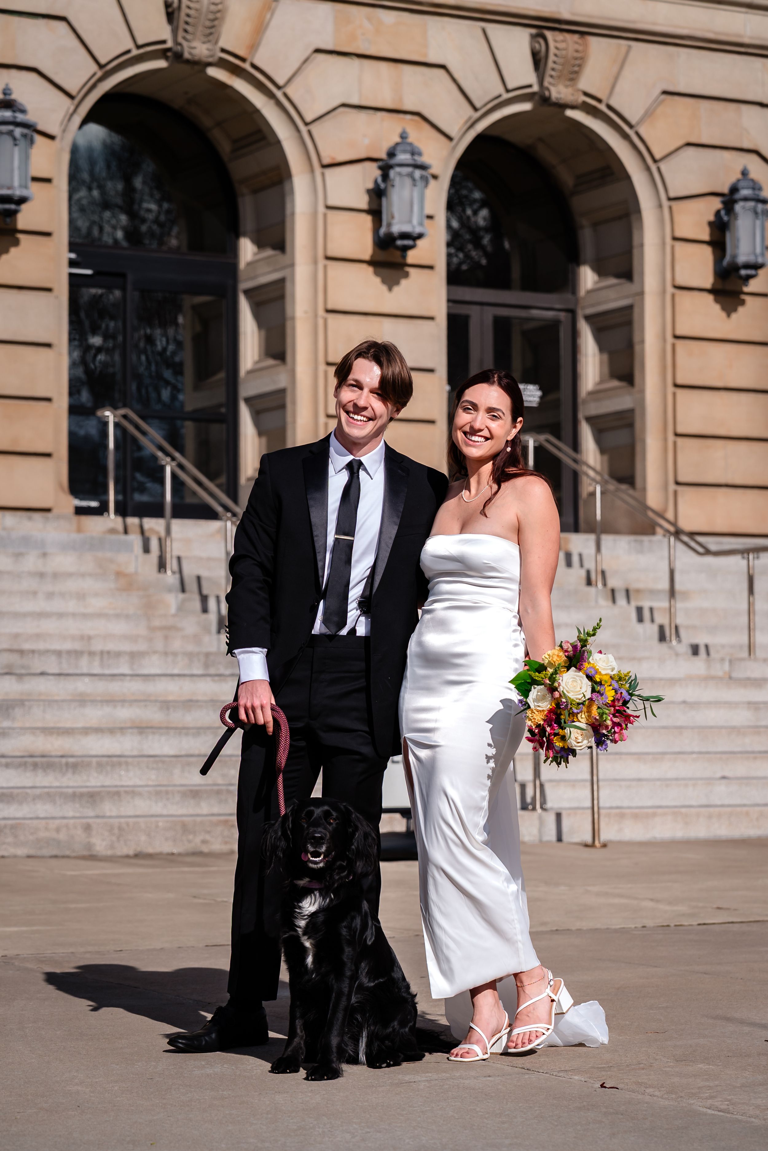 Couple in formal attire sitting on stone steps of historic building exterior.