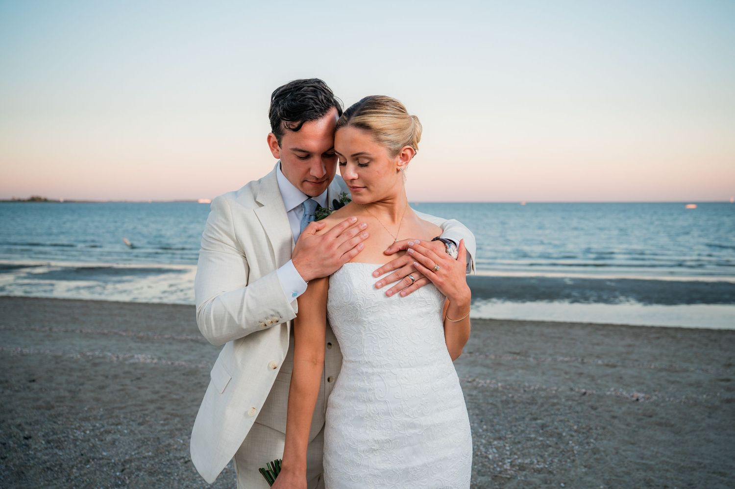 A romantic couple embraces on a beach at sunset while wearing white wedding attire.