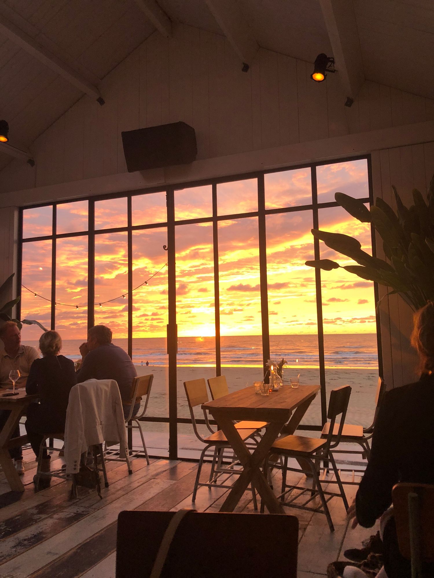 Diners enjoying a stunning fiery orange sunset ocean view through large windows of a coastal restaurant.