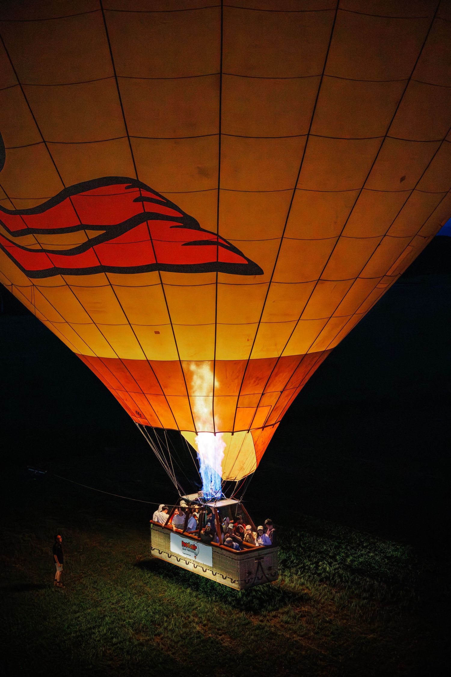 Hot air balloon glowing orange at night with flame burner illuminating passengers in basket on dark field. Cairns, Mareeba, Atherton, Aus