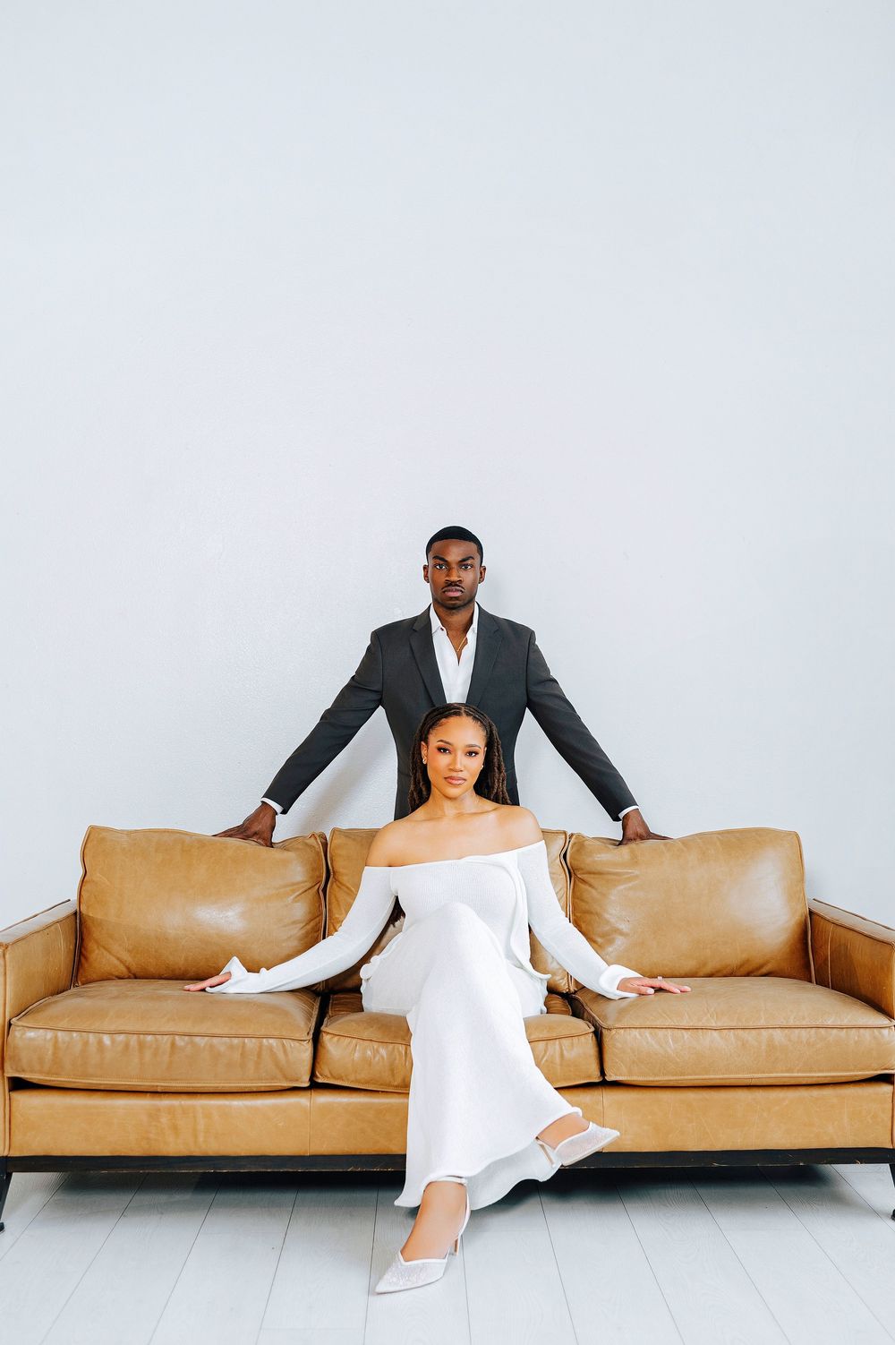 Elegant couple posing with a tan leather sofa, man in dark suit standing behind woman in white dress.