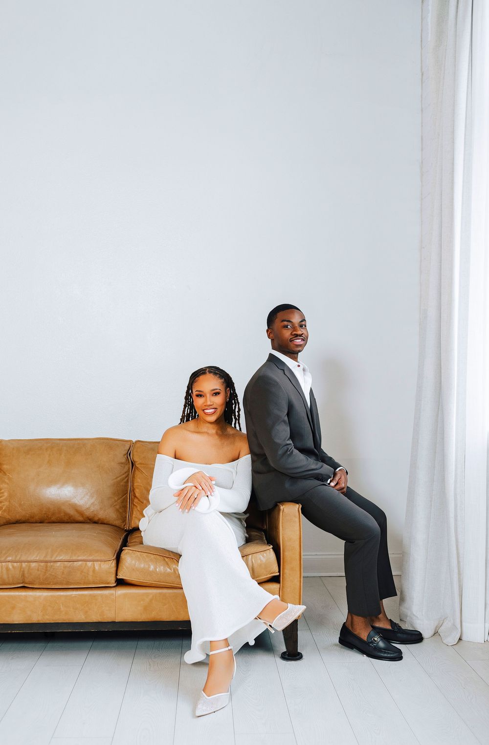 Elegant couple in white dress and gray suit posing on a tan leather couch in a bright minimalist studio.