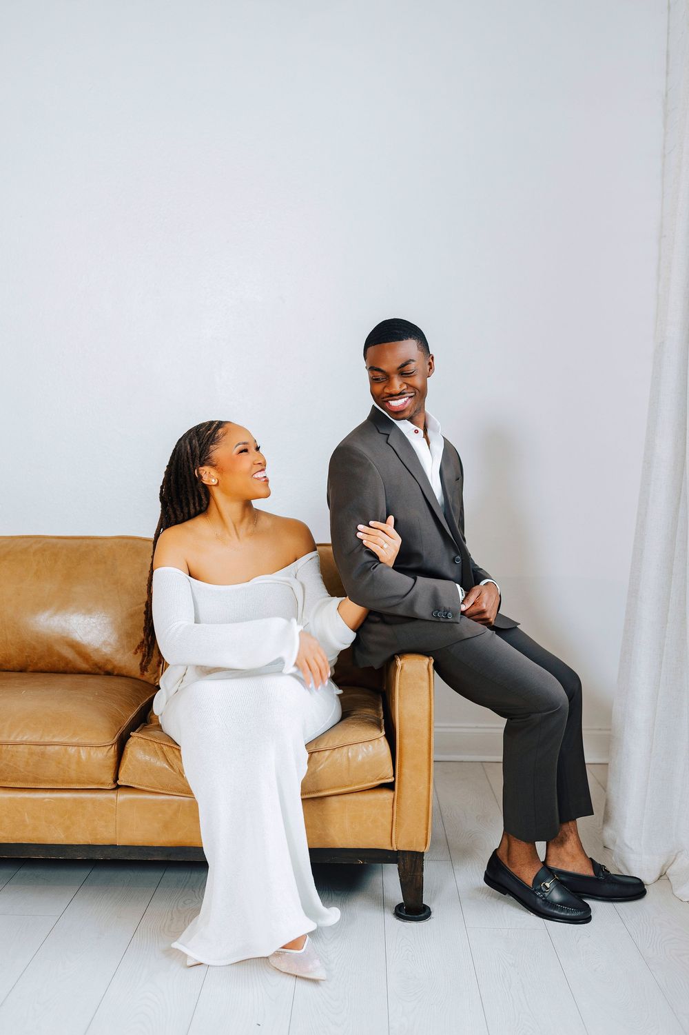 A joyful Black couple in elegant white dress and dark suit sitting together on a tan leather couch, smiling.