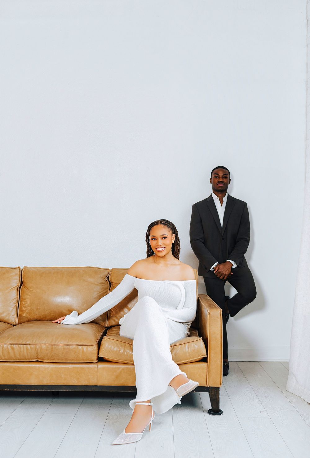 Elegant couple in white outfit and black suit posing with a tan leather sofa against a light gray wall.