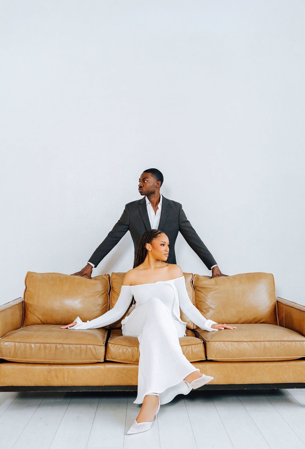 A stylish Black couple poses with a tan leather sofa, woman in white dress seated, man in gray suit standing behind.