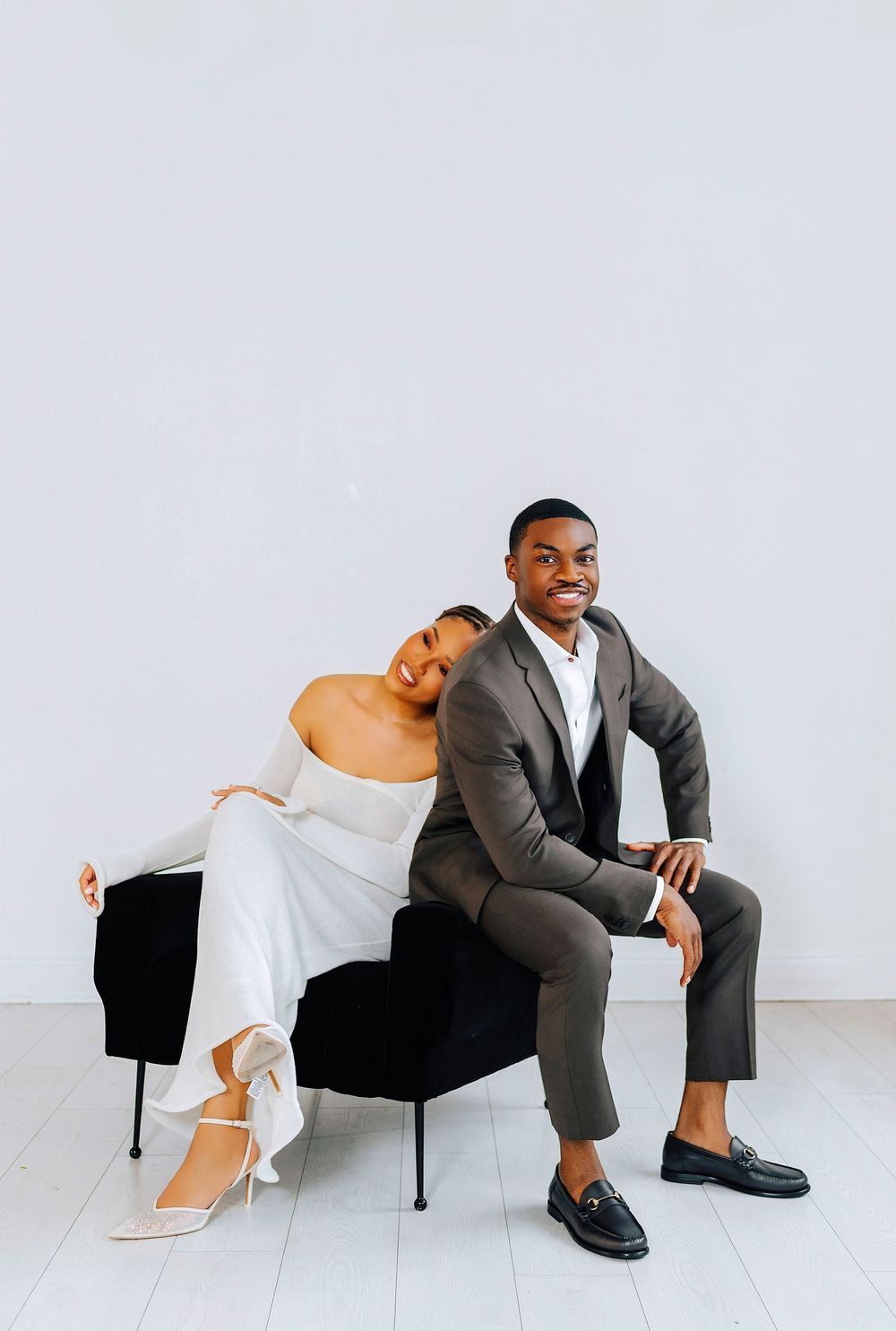 Elegant couple in wedding attire sitting on a black chair, woman in white gown leaning on man in brown suit.