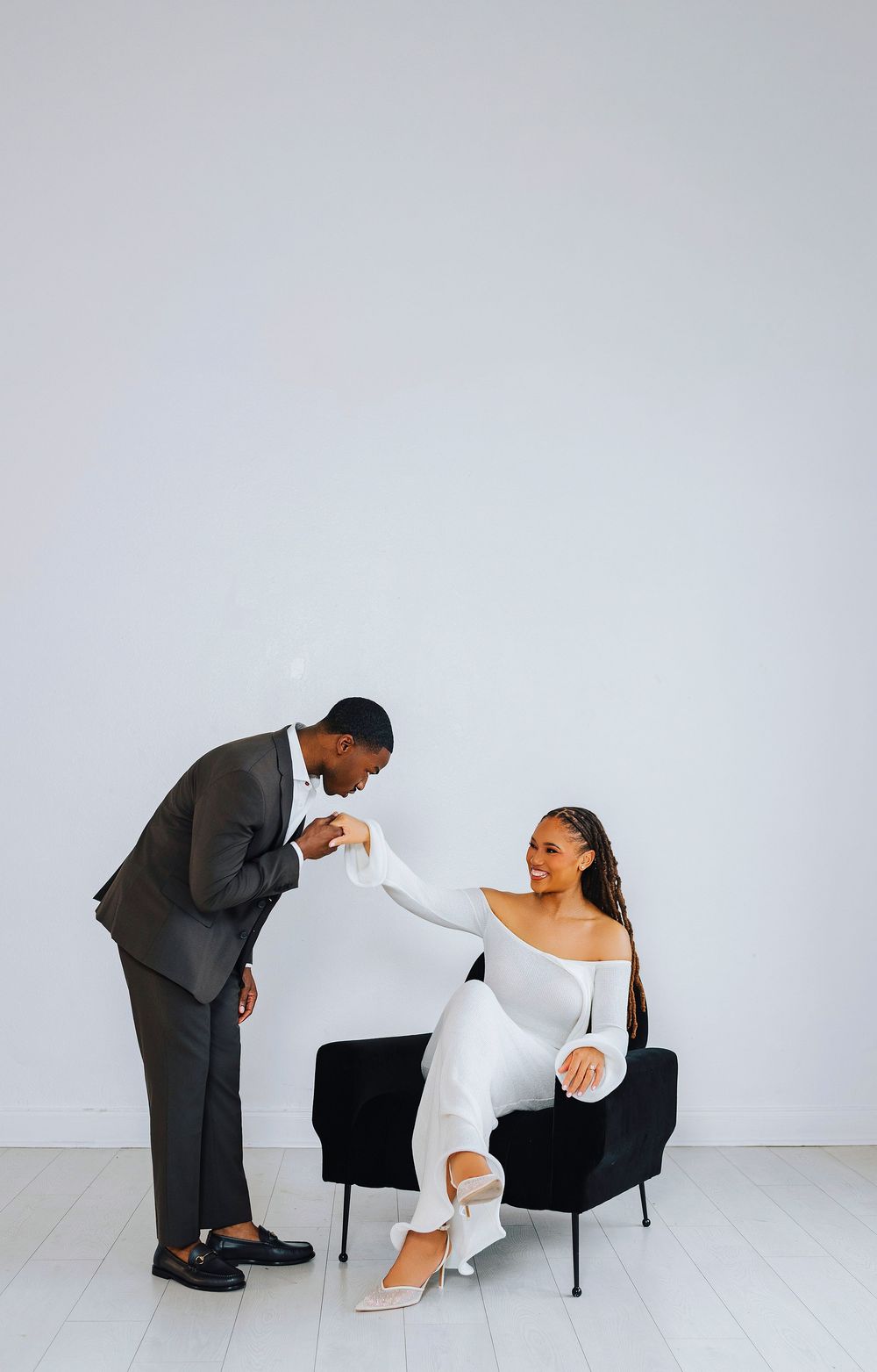 Man in gray suit kissing woman's hand as she sits in black chair wearing white outfit, romantic pose.