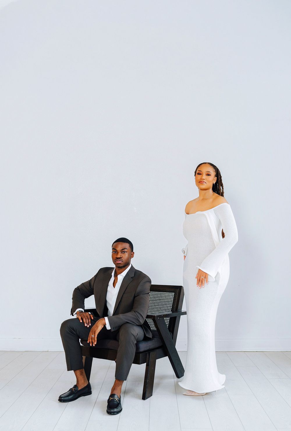 Couple posing with man seated in black chair in gray suit, woman standing in white off-shoulder gown beside him.