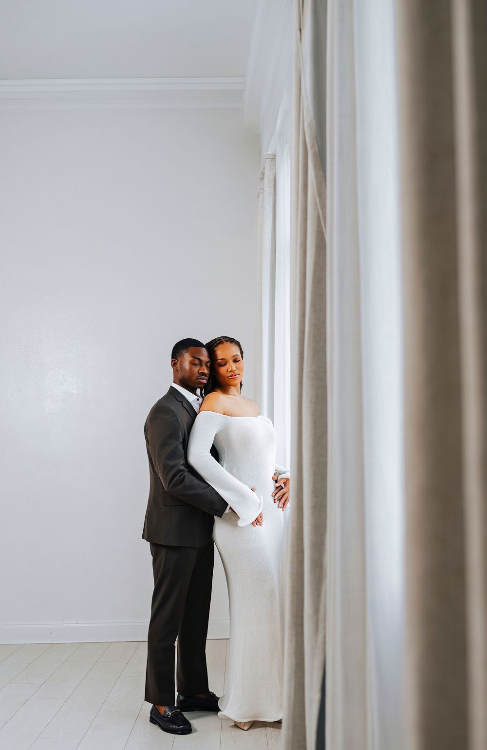 A Black couple embraces near a curtained window, woman in white off-shoulder gown, man in dark suit holding her close.