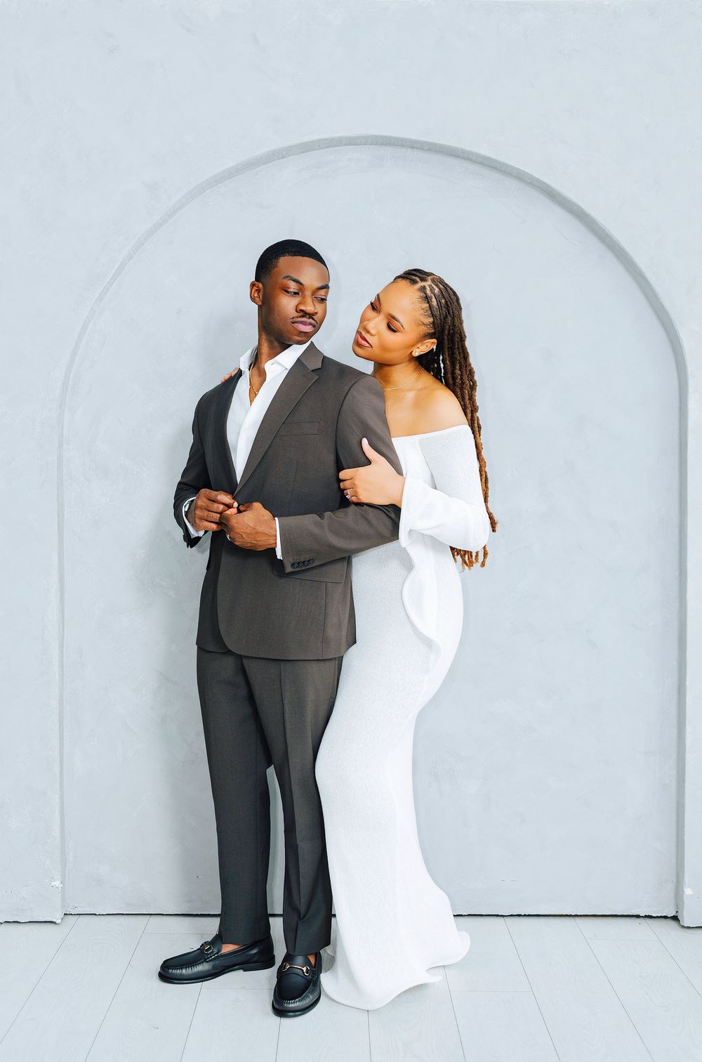 Elegant couple posing by arch, man in gray suit, woman in white off-shoulder gown embracing him.
