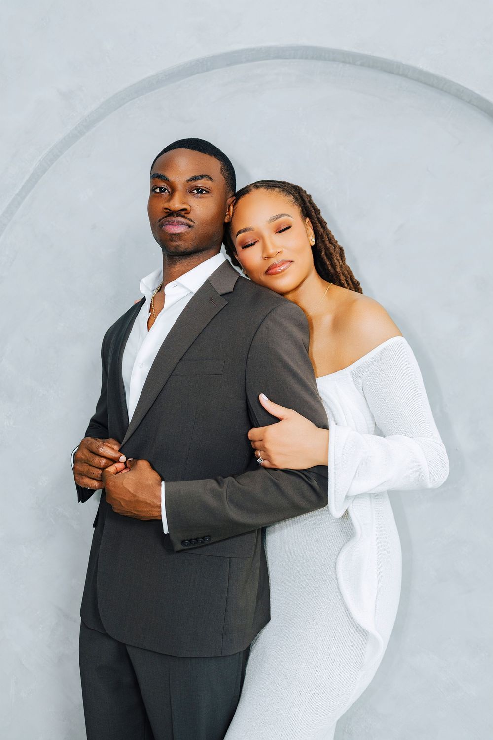 Stylish couple in formal wear, woman in white dress resting head on mans shoulder against gray wall.